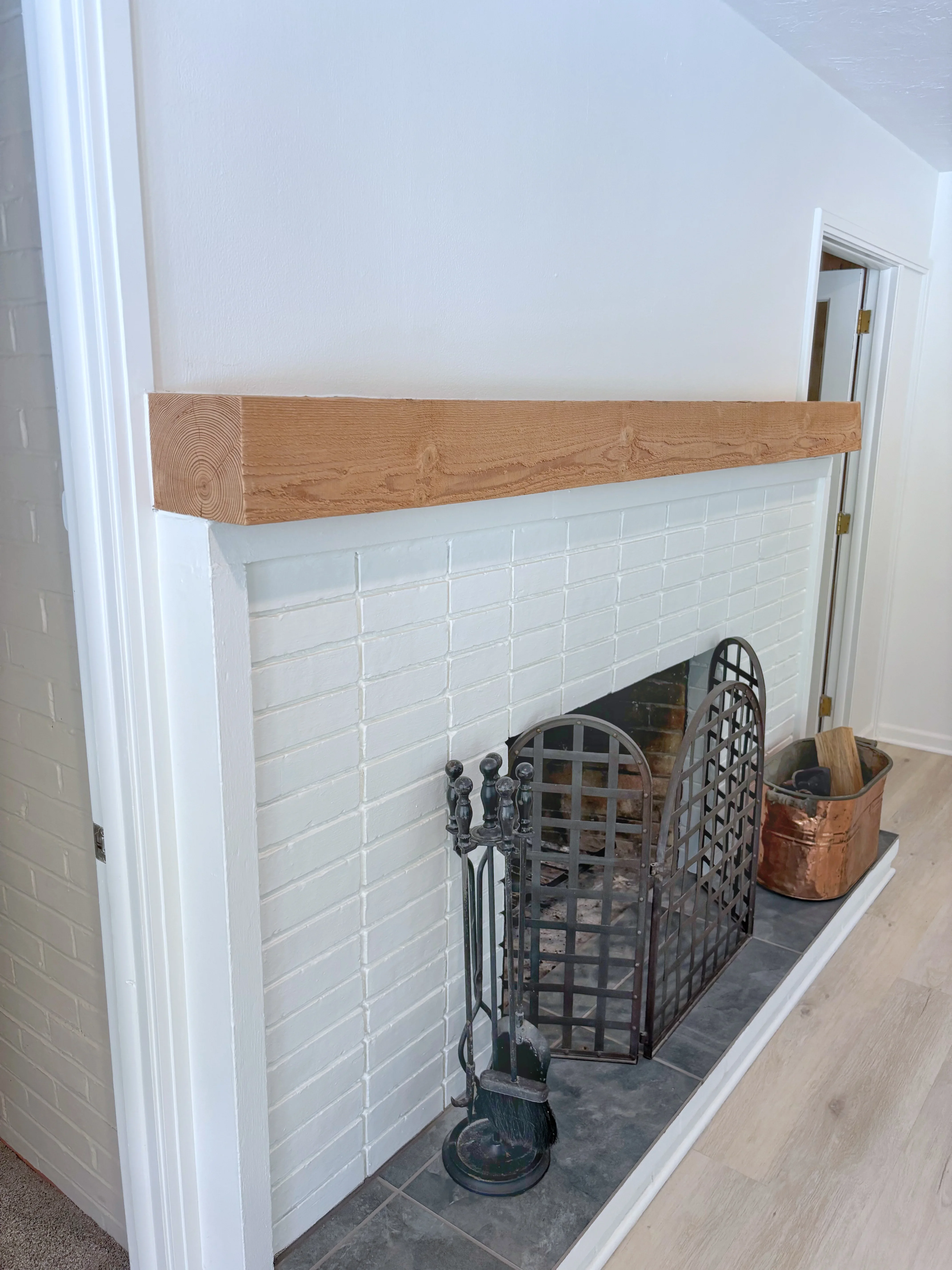 Oak wood mantel over white subway tile fireplace surround with black hearth and fireplace tools