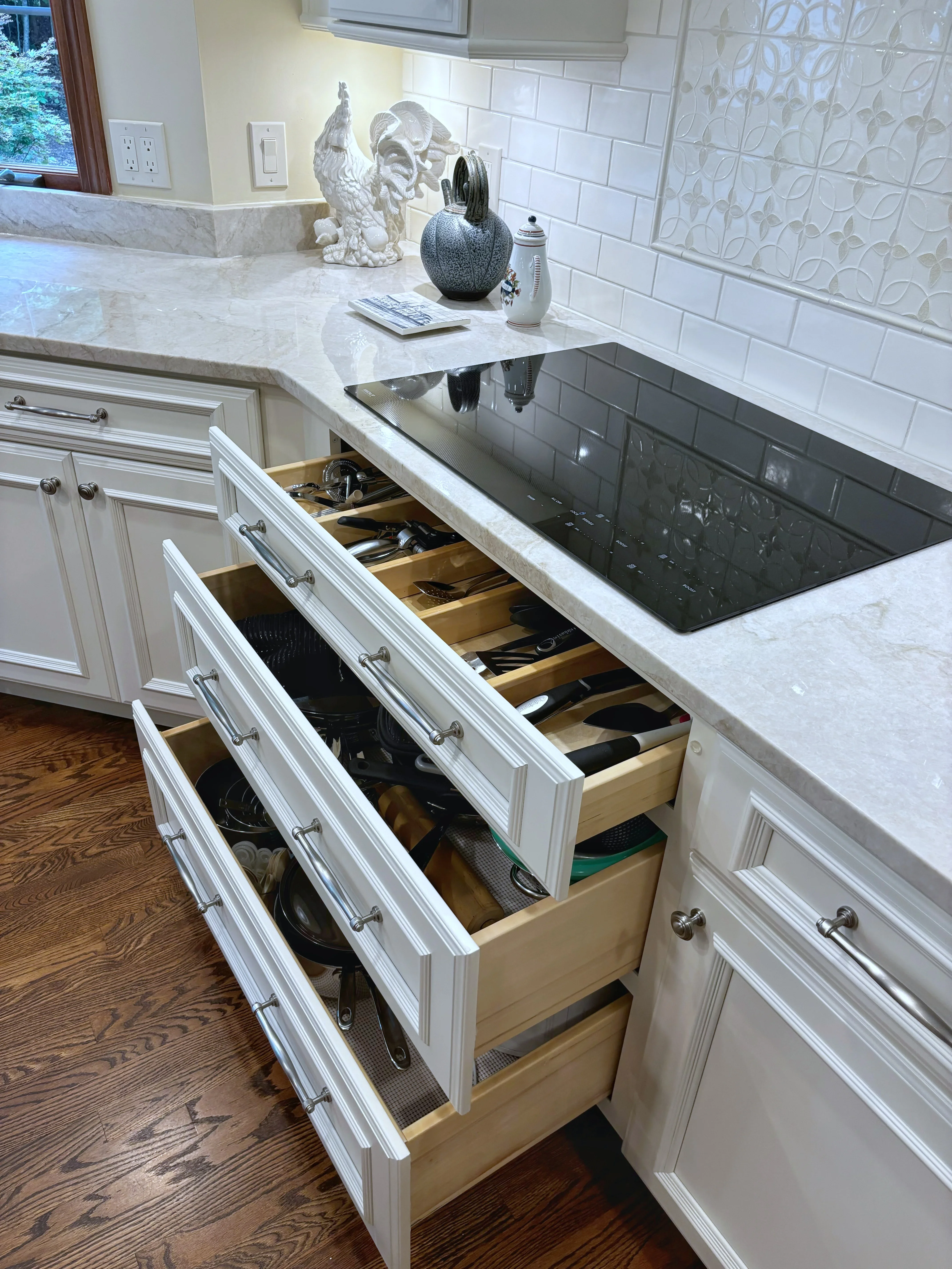 White kitchen cabinetry with marble countertops, open drawers showing wood interiors and cookware storage, black induction cooktop