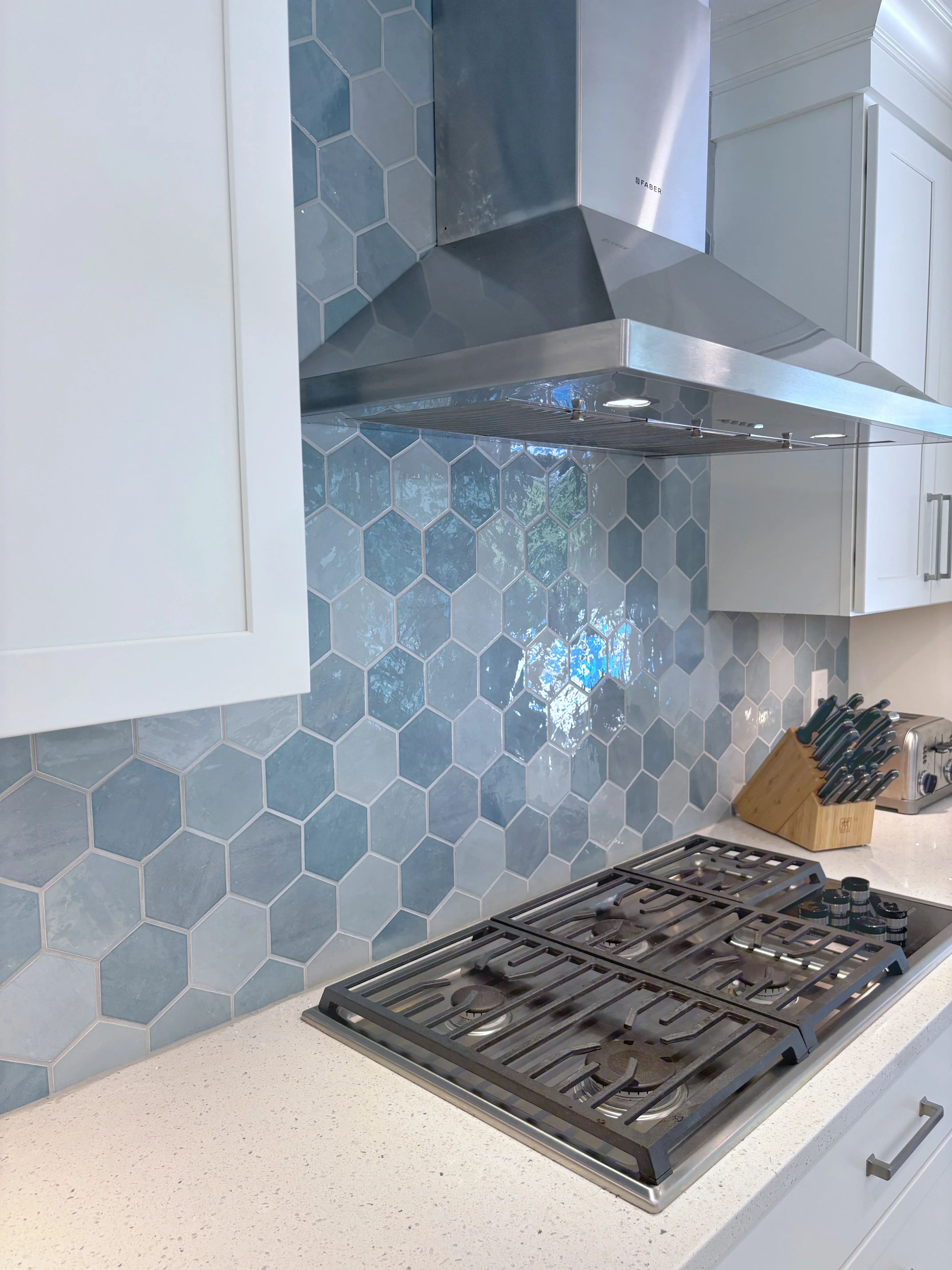 Blue hexagonal tile backsplash, stainless steel range hood, gas cooktop, white cabinetry, quartz counters