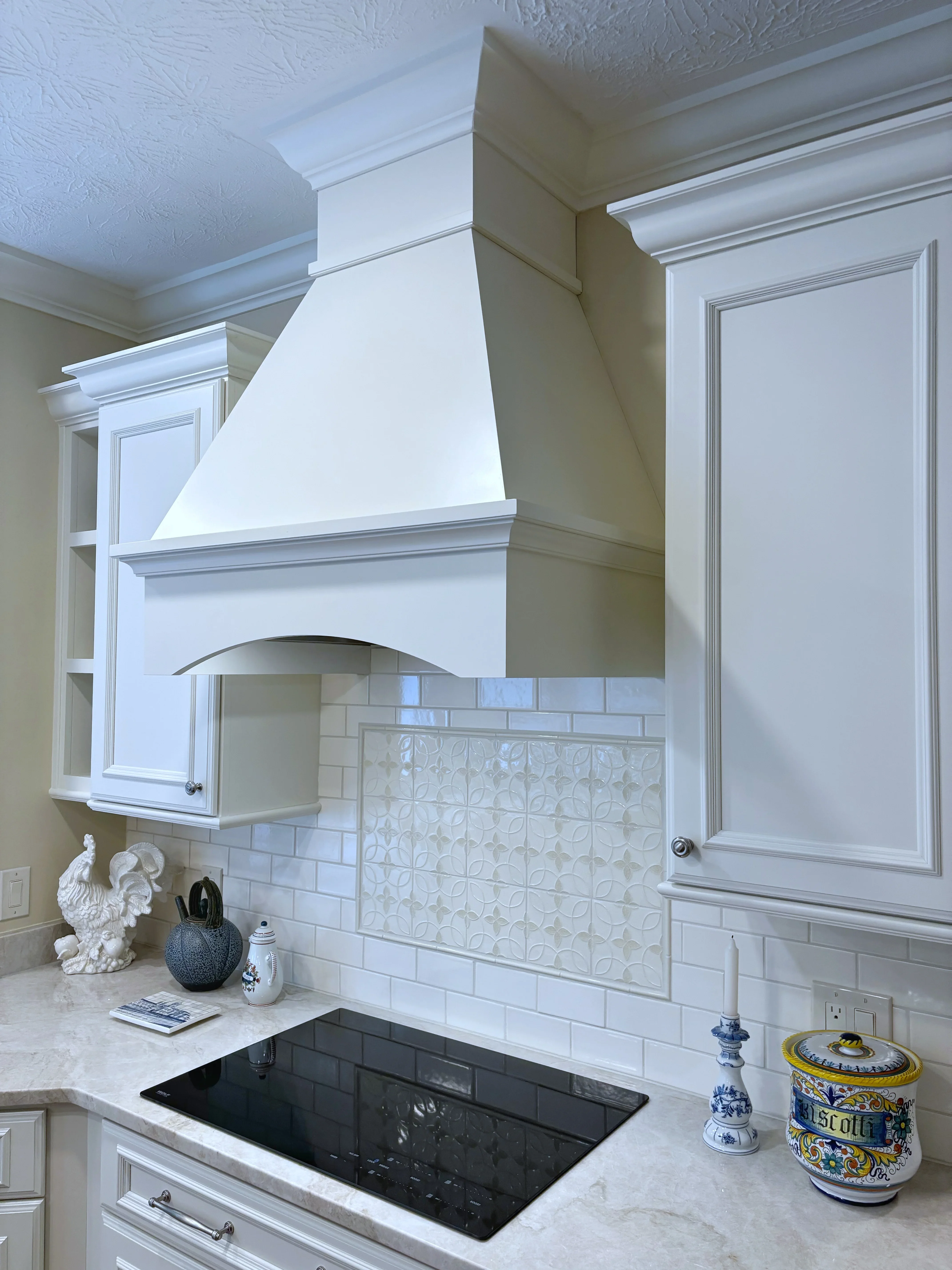 White custom range hood with curved design, subway tile and textured backsplash, marble countertop, black induction cooktop