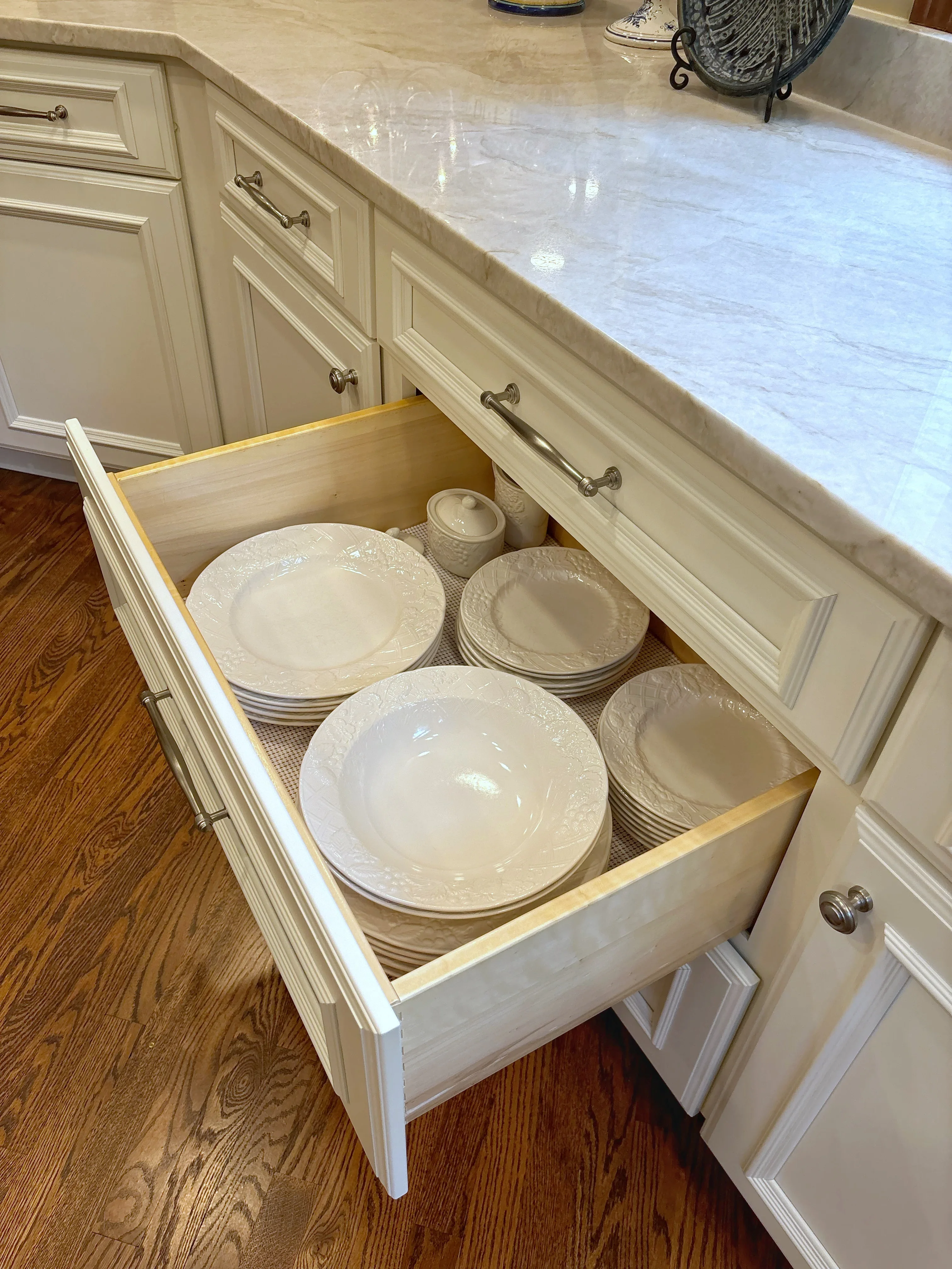Cream kitchen cabinet drawer open showing marble countertop, white dinnerware storage, and brushed nickel hardware